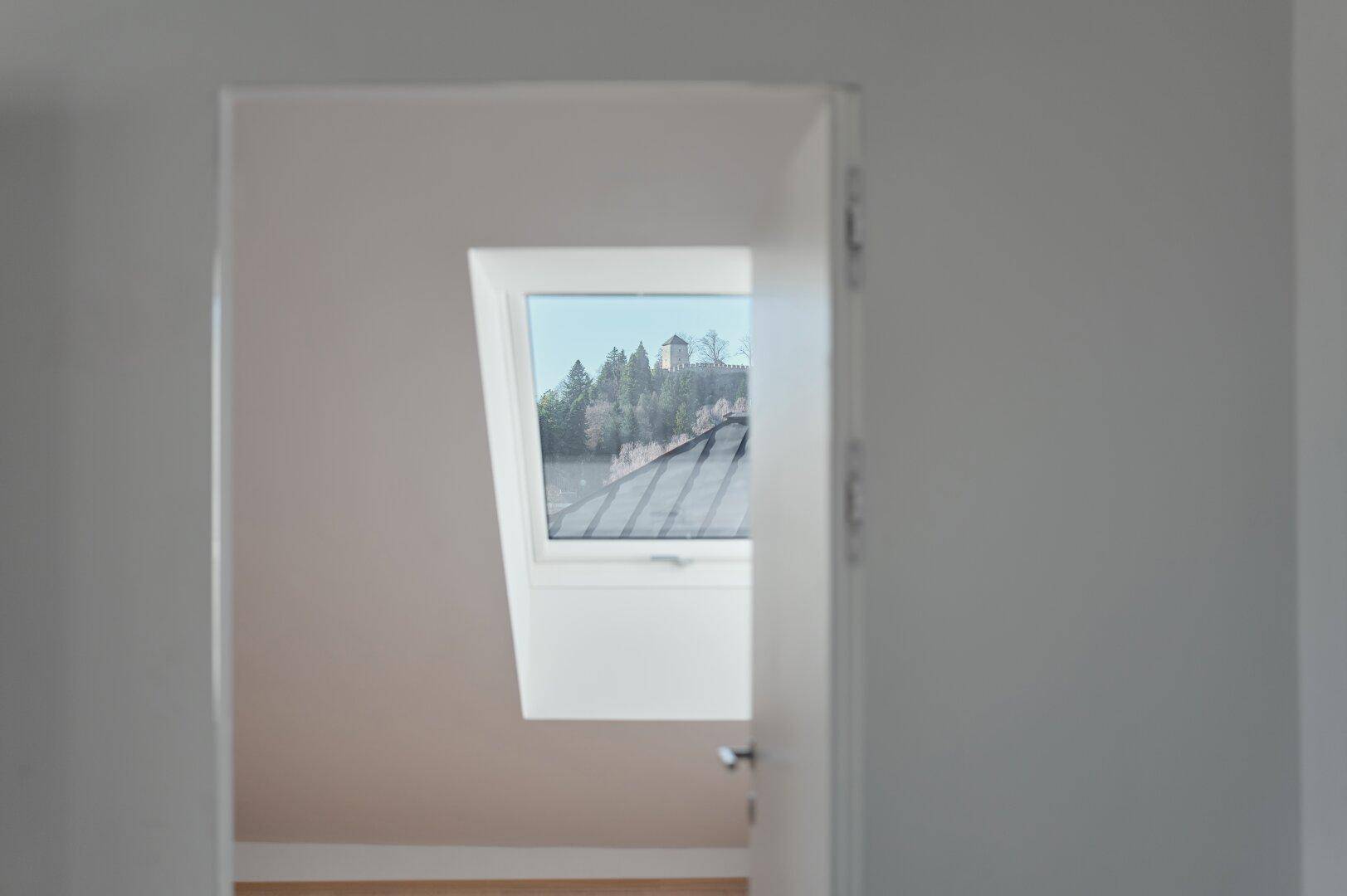 Blick durch eine offene Tür auf einen Raum mit Dachfenster und Blick auf eine bewaldete Anhöhe.