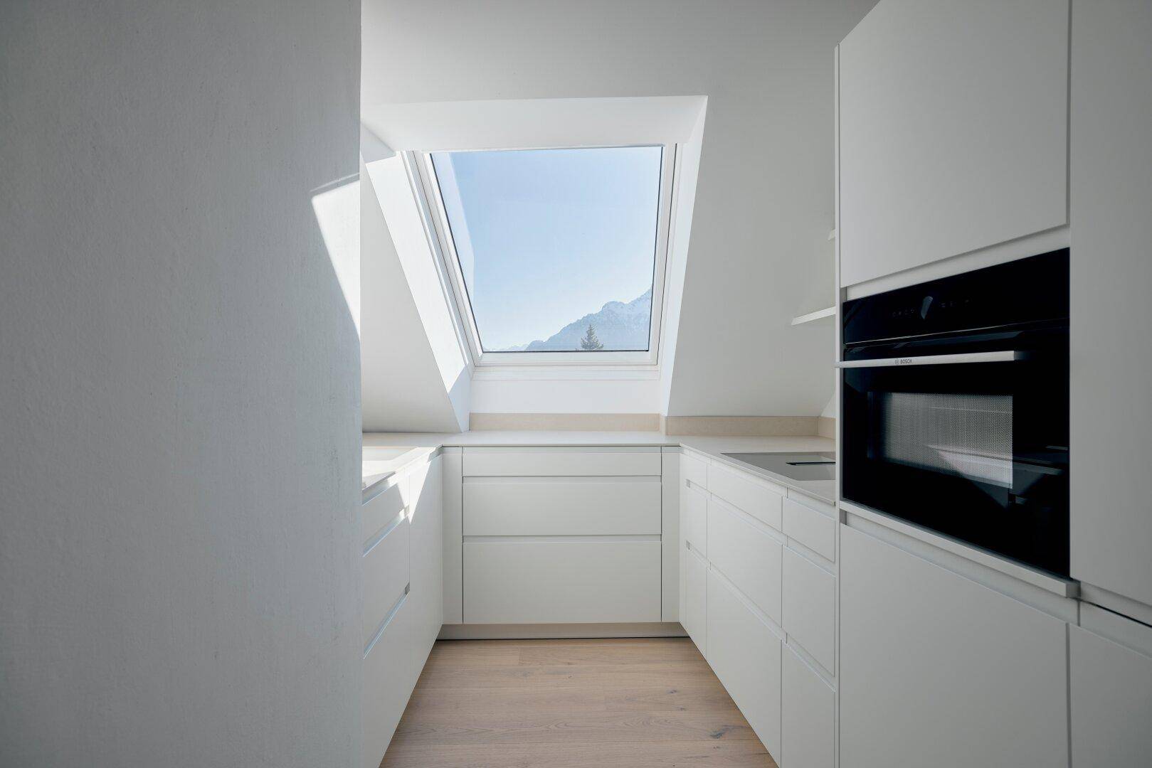 Moderne weiße Küche im Dachgeschoss mit Einbaugeräten, Dachfenster und Bergblick.