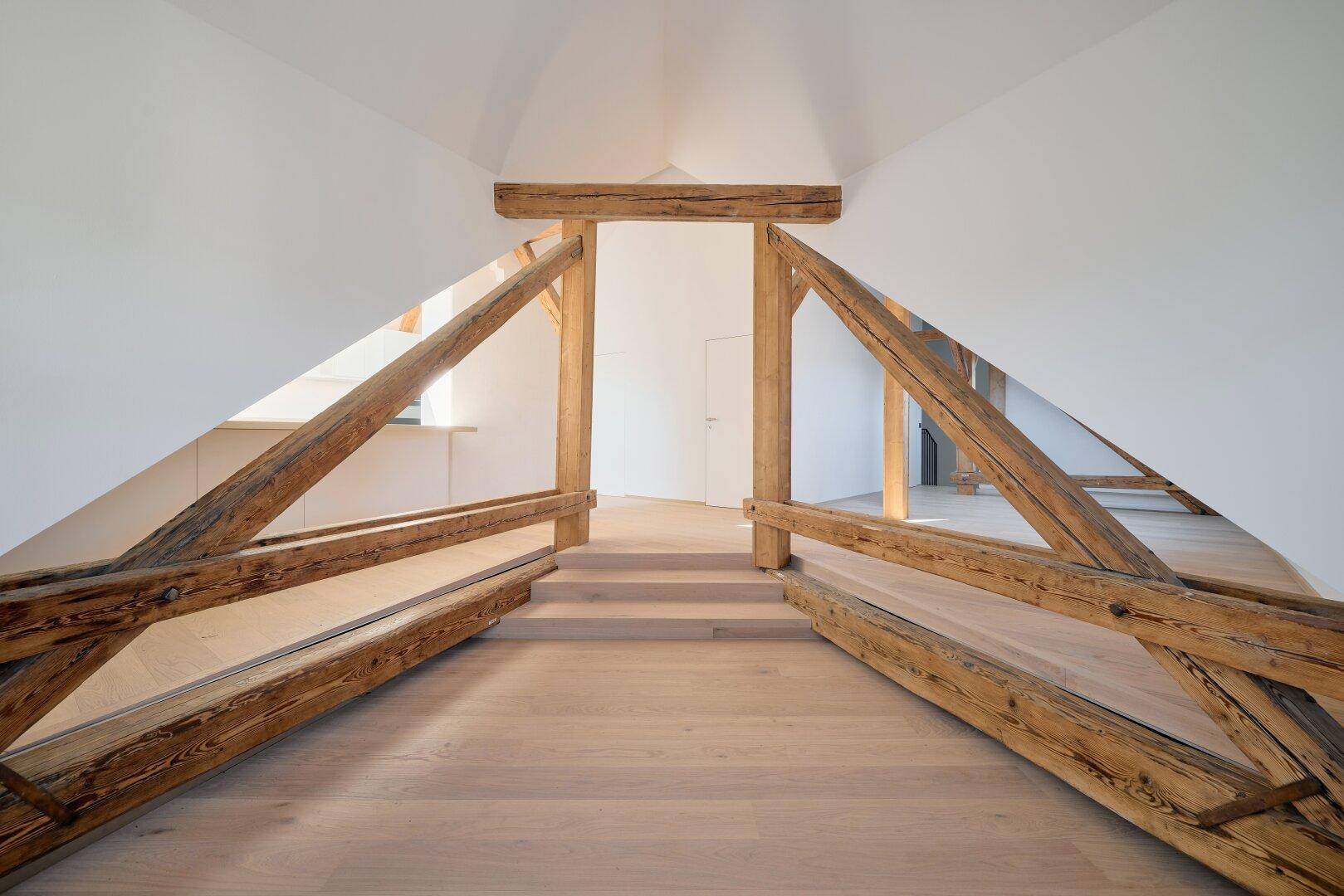 Offener Dachbodenbereich mit markanten Holzbalken, hellem Holzboden und schrägen Wänden.