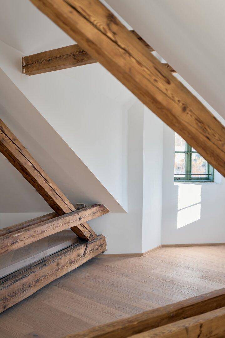 Heller Dachbodenraum mit sichtbaren Holzbalken, hellem Holzboden und einem Fenster mit grünen Rahmen.