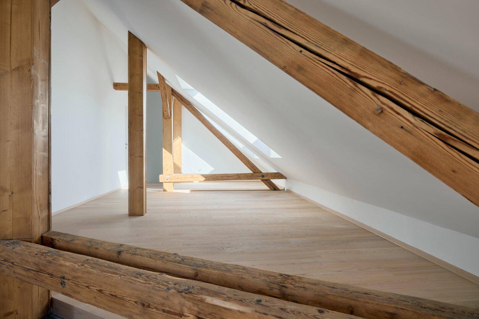 Leerer Dachbodenraum mit schrägen Wänden, Holzbalken und hellem Holzboden, gut beleuchtet.