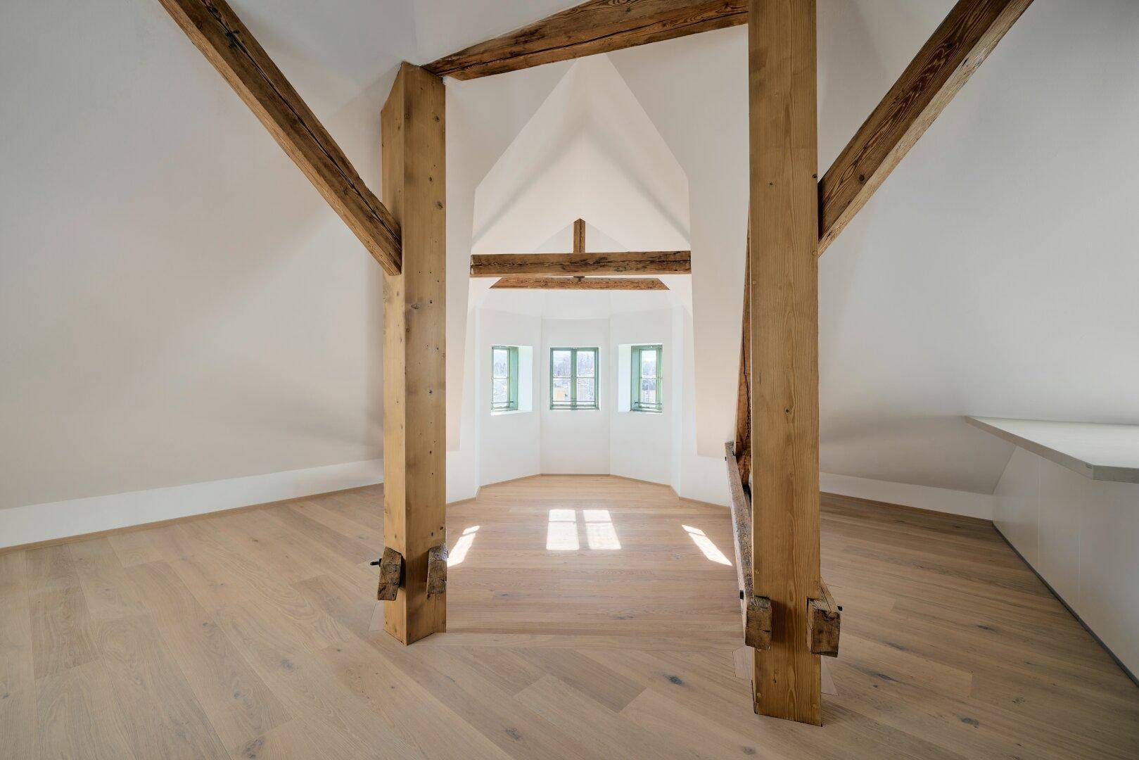 Geräumiger Dachbodenraum mit sichtbaren Holzbalken, hellem Parkettboden und kleinen Fenstern.