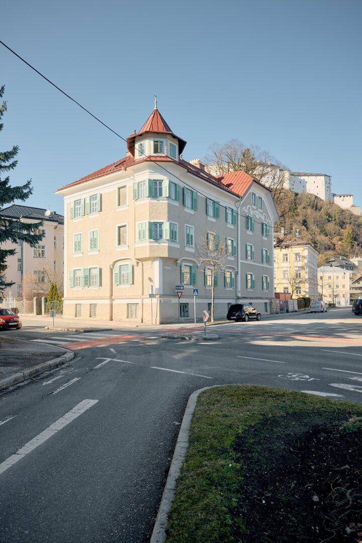 Historisches Gebäude mit markantem Turm und roten Dachziegeln, im Hintergrund eine Burg.