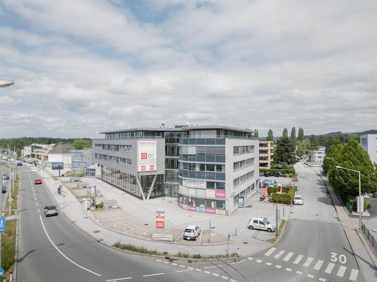 Modernes Geschäftsgebäude an einer Kreuzung mit umliegenden Straßen und Parkplätzen unter bewölktem Himmel.