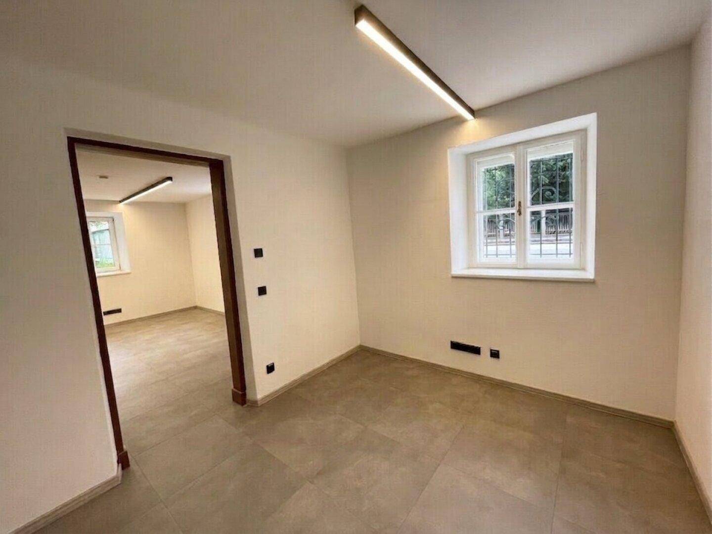 Heller, leerer Raum mit einem Fenster, modernen Deckenleuchten und einem Fliesenboden.