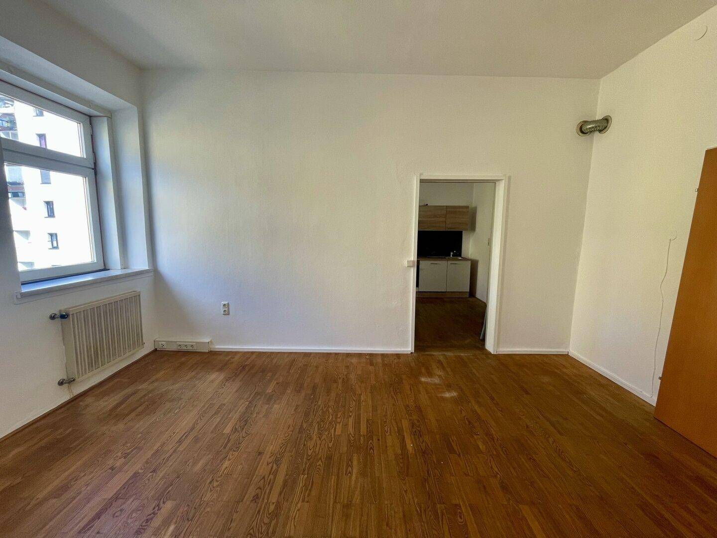Geräumiges, helles Zimmer mit Holzboden und Blick auf die umliegenden Gebäude.