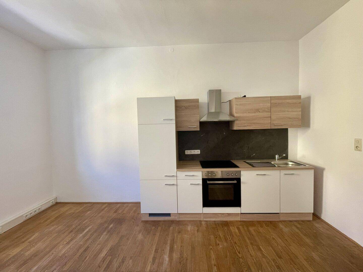 Moderne Küchenzeile mit hellen Fronten, integriertem Backofen und Dunstabzugshaube in einem leeren Raum.