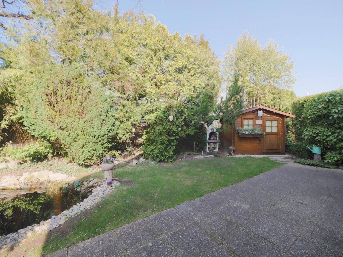 Idyllischer Gartenbereich mit Teich, Gartenhaus und Grillplatz unter hohen Bäumen.