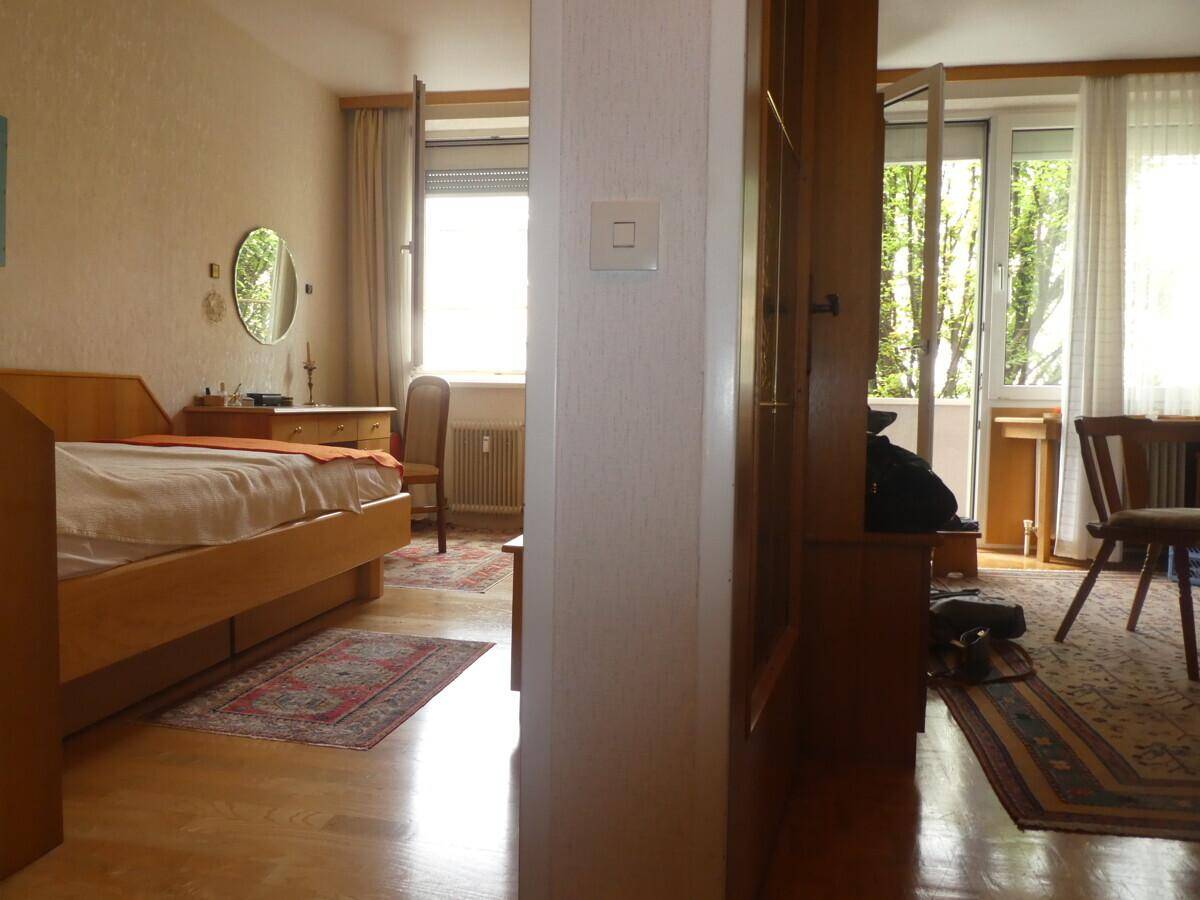 Schlafzimmer mit Holzbett und Zugang zu einem weiteren Raum, hell durch große Fenster.