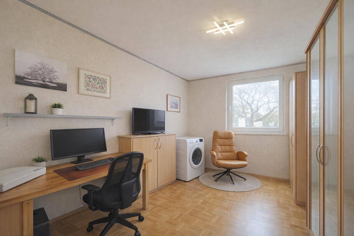 Arbeitszimmer mit Schreibtisch, Computer und Waschmaschine, hell durch ein großes Fenster.