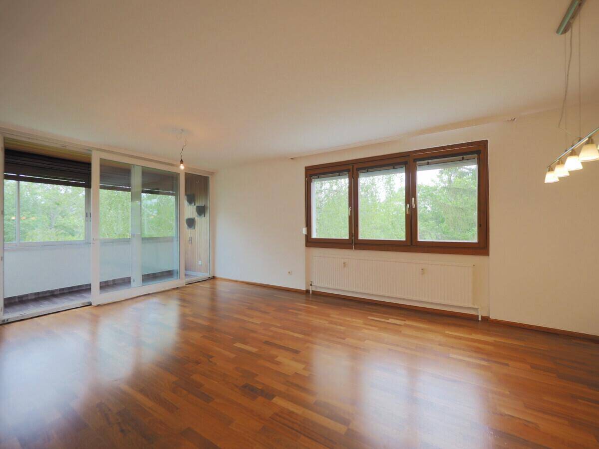 Geräumiges Wohnzimmer mit Holzboden, großen Fenstern und Zugang zu einem Balkon.