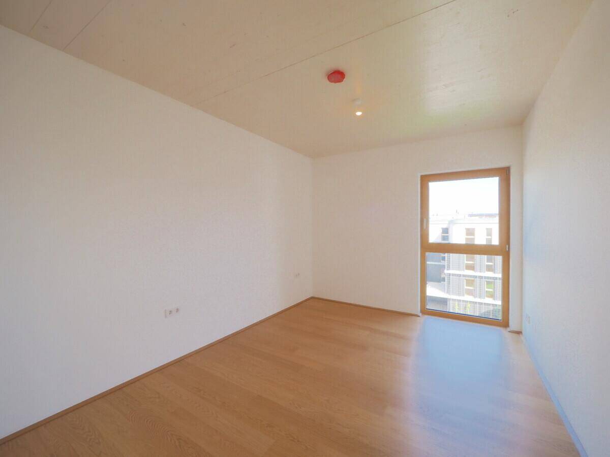 Helles Schlafzimmer mit Parkettboden und großem Fenster, das viel Tageslicht hereinlässt.