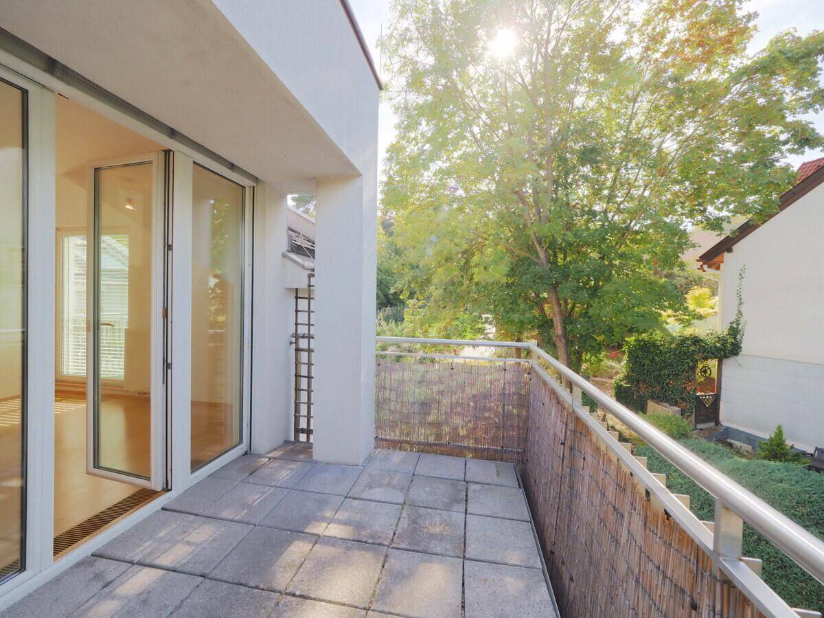 Sonniger Balkon mit Steinplatten, Metallgeländer und Blick ins Grüne mit Bäumen und Häusern.