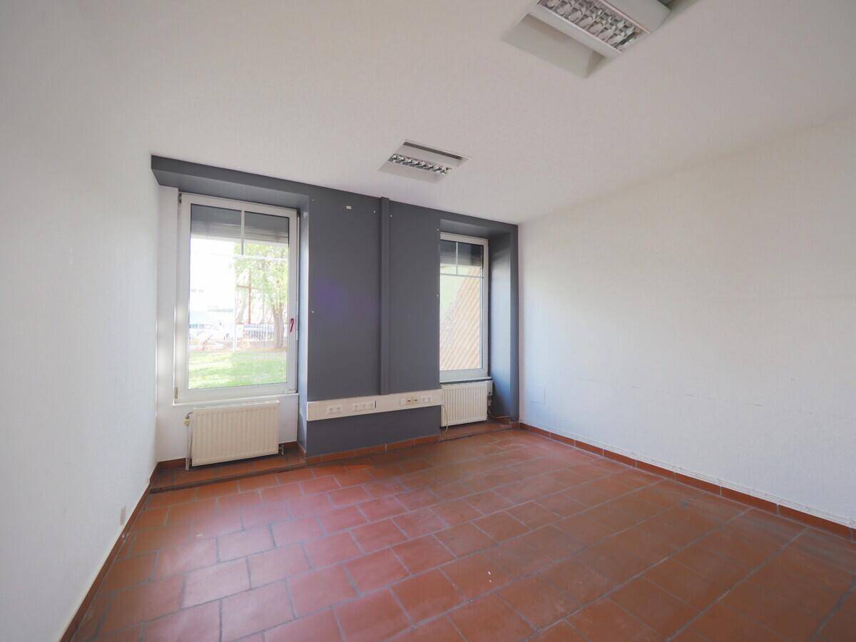 Büroraum mit Terrakottafliesen, weißen Wänden und zwei Fenstern mit dunklen Akzenten.