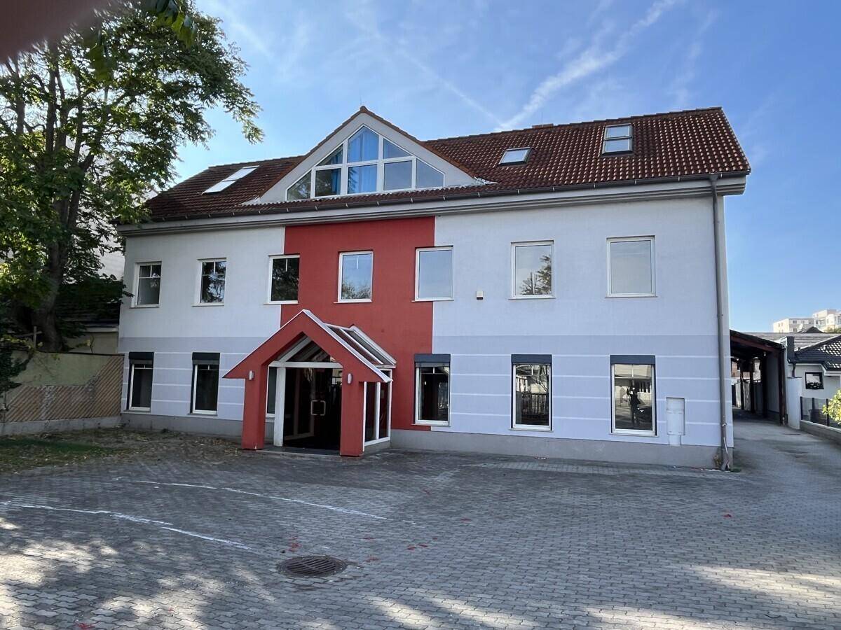 Seitenansicht des Gebäudes mit rotem Akzent und gepflastertem Vorplatz unter blauem Himmel.