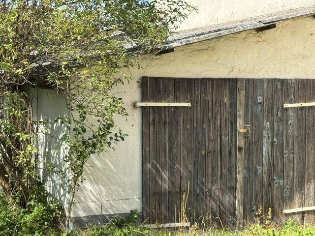 Verwittertes Holztor an einer Außenwand, umgeben von Vegetation, was auf einen ländlichen Stil hindeutet.