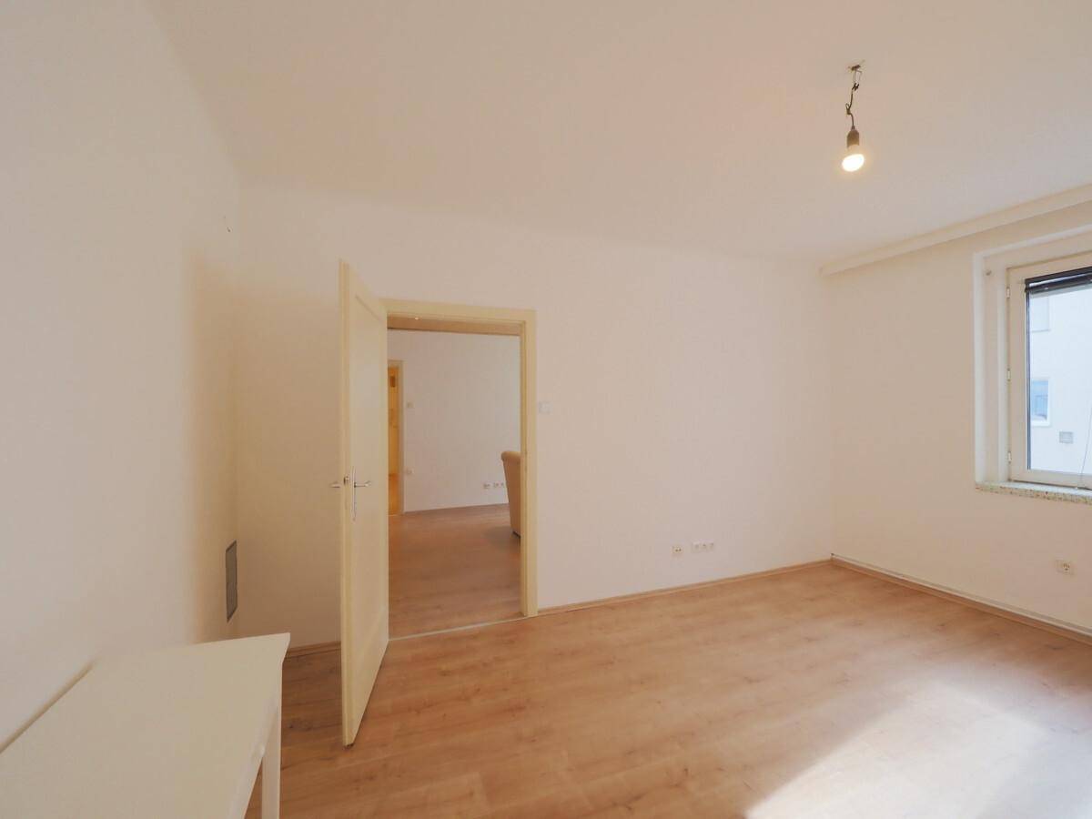 Unmöblierter Raum mit Laminatboden und Fenster, das Tageslicht hereinlässt, mit Zugang zu einem weiteren Zimmer.