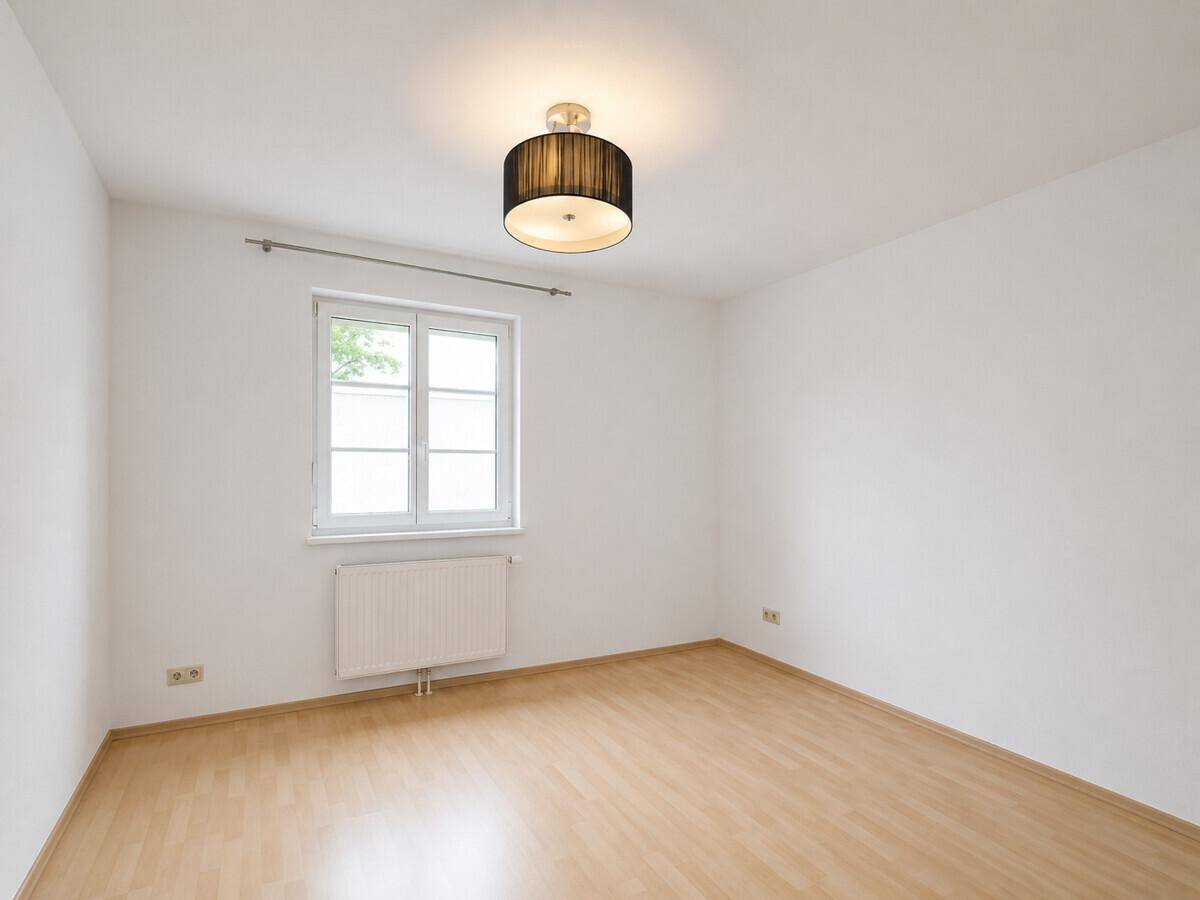 Helles, leeres Zimmer mit Fenster, Holzboden und moderner Deckenleuchte.