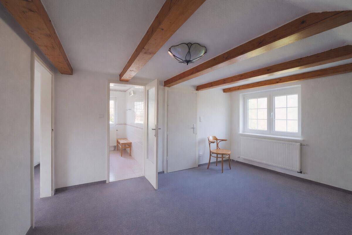 Helles Dachzimmer mit Holzbalken, Teppichboden und Zugang zu einem weiteren Raum.