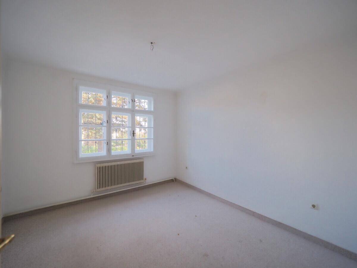 Leeres Zimmer mit Teppichboden, großem Fenster und Heizkörper, bereit zur individuellen Gestaltung.