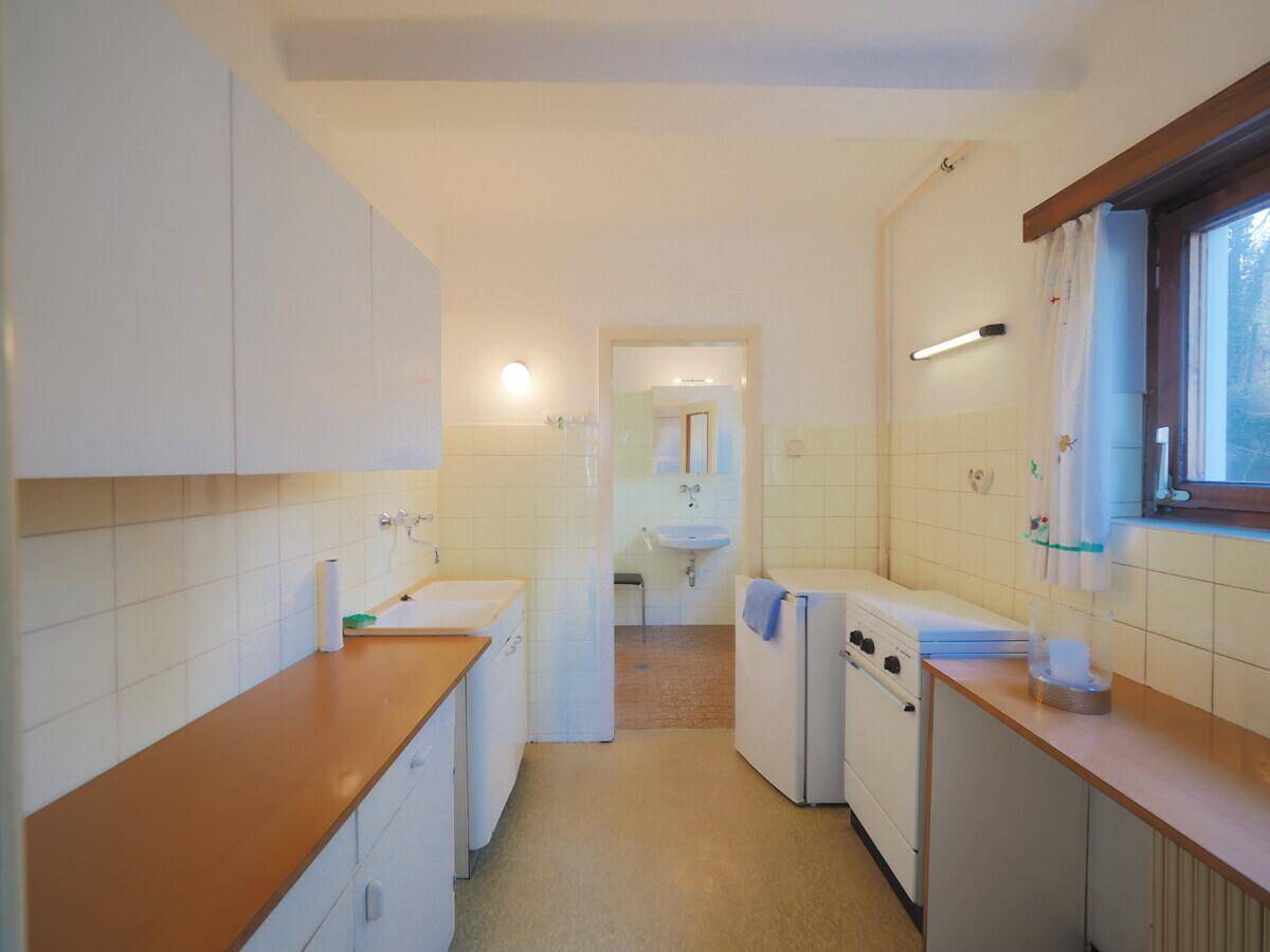 Schmale Küche mit einfachen weißen Schränken, gelben Fliesen und Blick in ein angrenzendes Badezimmer.