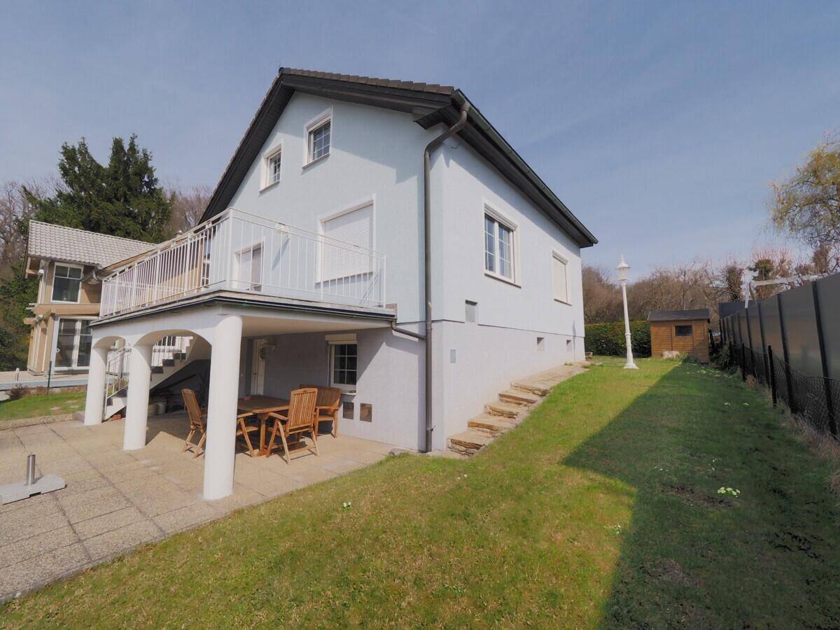 Hausfassade mit überdachter Terrasse, Garten und einem kleinen Gartenhaus im Hintergrund.