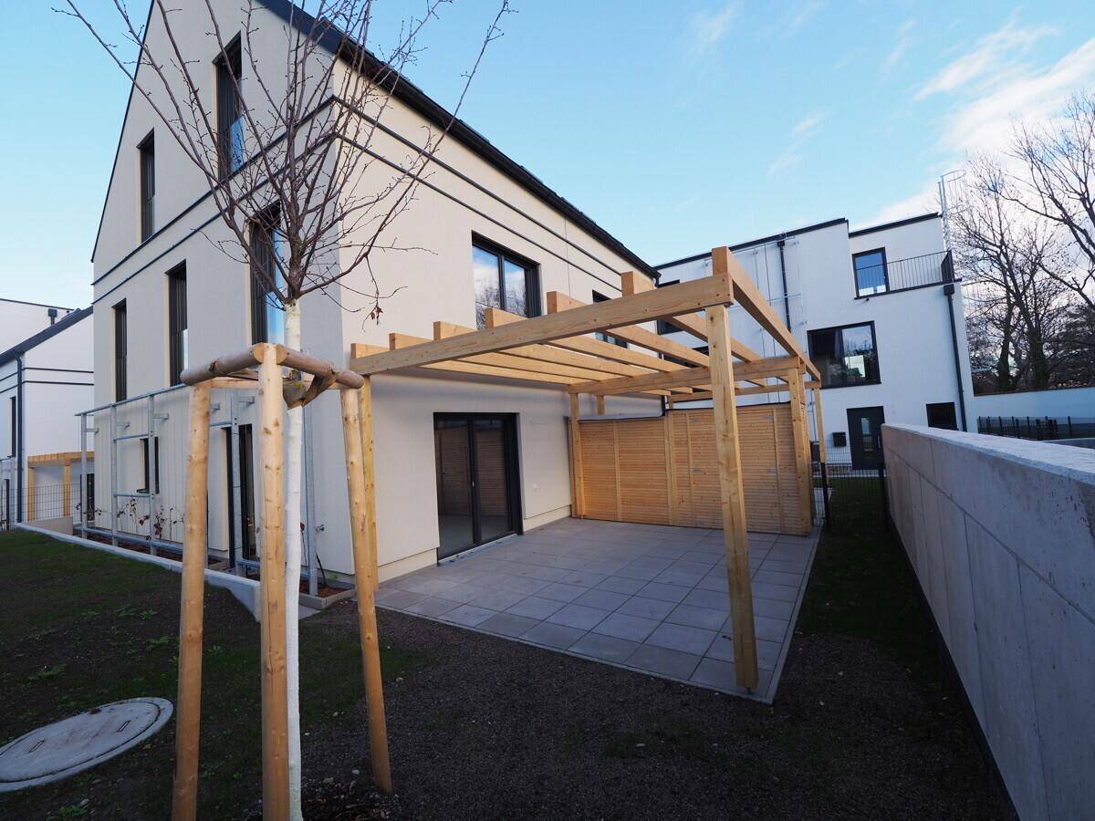 Terrasse mit Holzpergola und jungem Baum, umgeben von einer niedrigen Mauer und Rasenfläche.