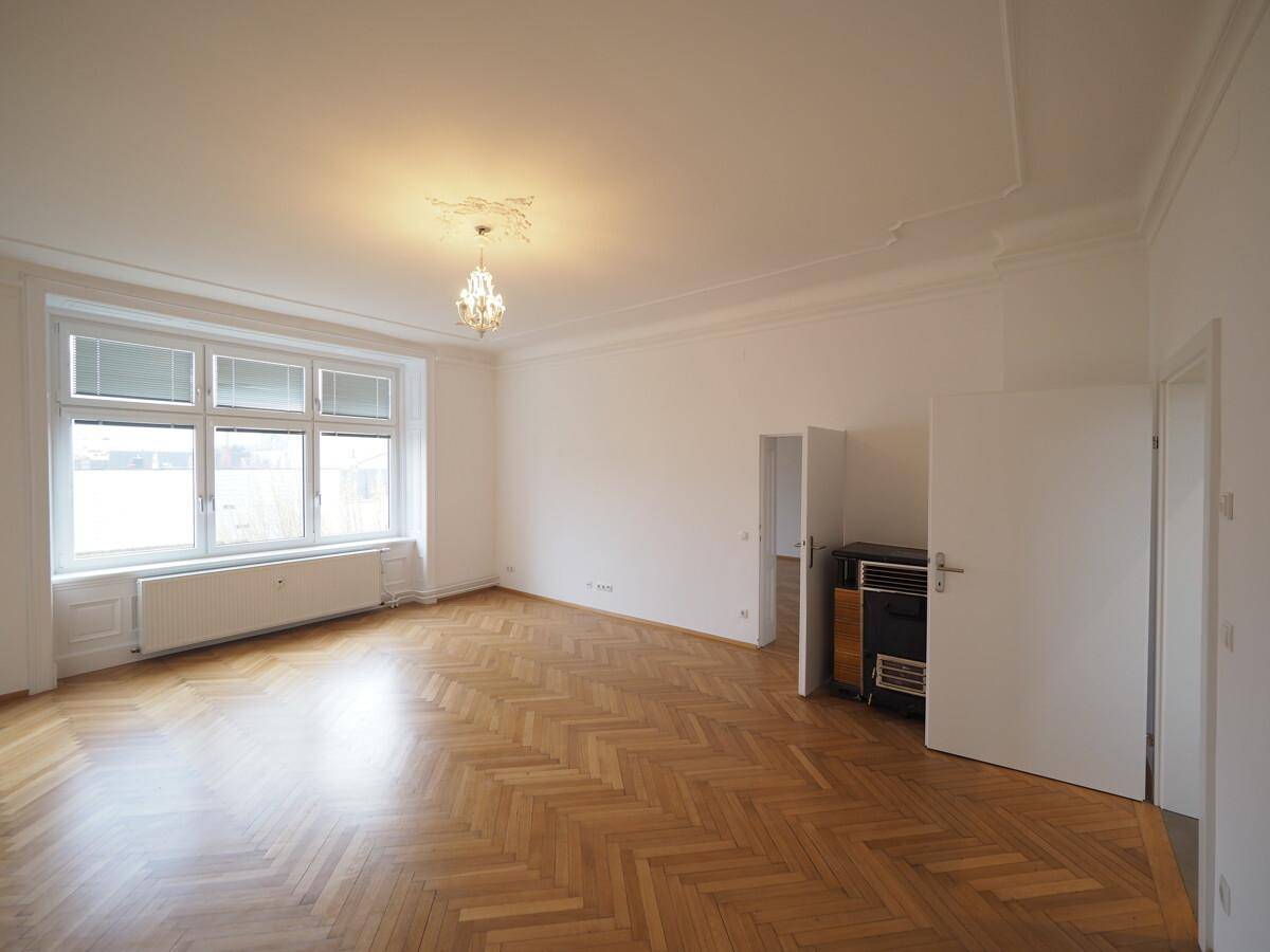 Geräumiges Wohnzimmer mit Fischgrätparkett, großen Fenstern und einem Kachelofen.