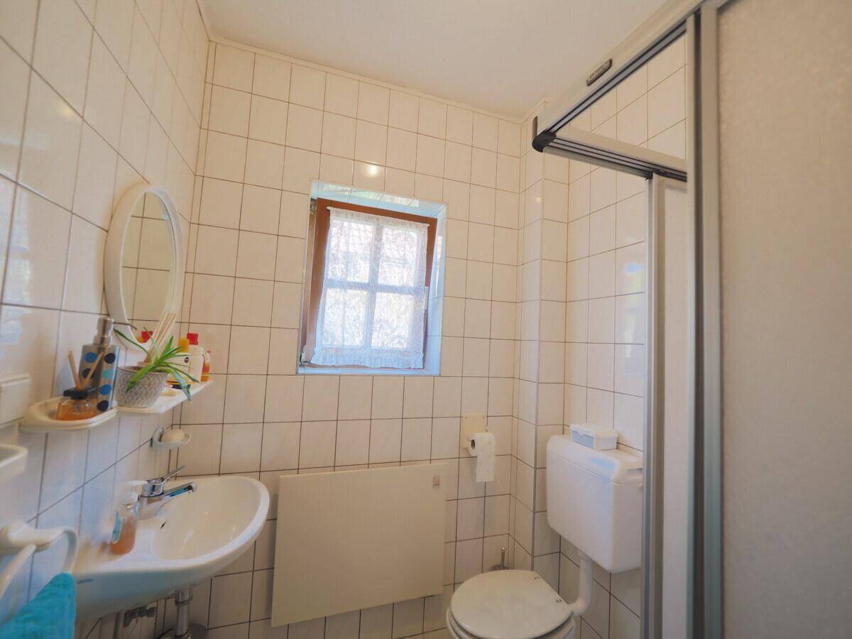 Kleines Badezimmer mit weißen Fliesen, Dusche, WC und einem Fenster für Tageslicht.