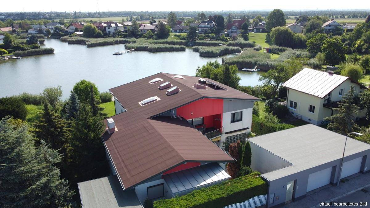 Vogelperspektive des Wohnhauses mit braunem Dach, umgeben von viel Grün und einem See im Hintergrund.
