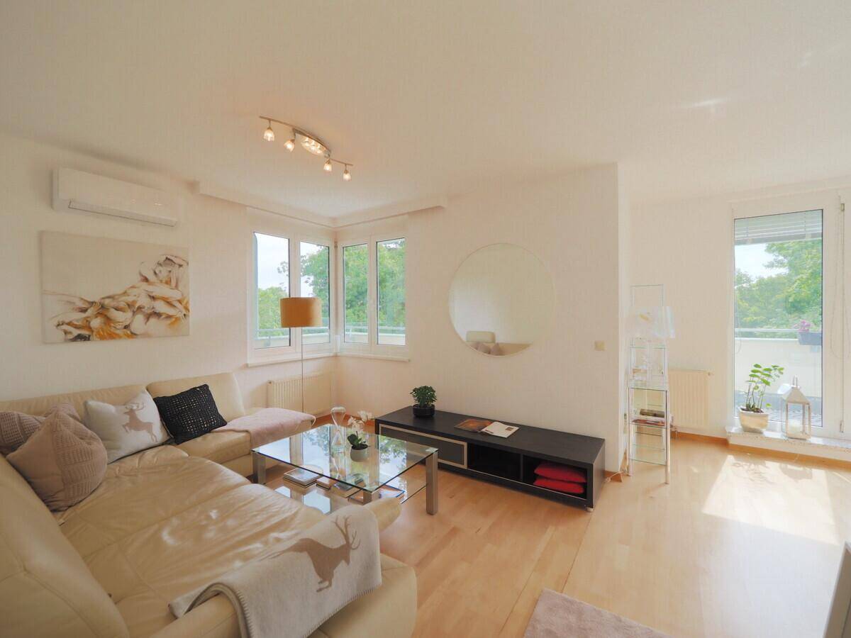 Heller Wohnbereich mit beigem Ecksofa, Holzboden und Blick ins Grüne durch große Fenster.