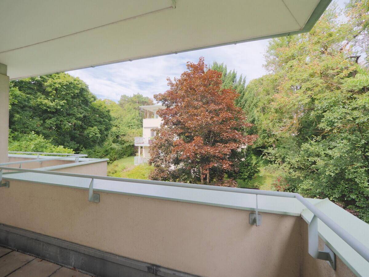 Blick vom Balkon auf die grüne Umgebung und ein weiteres Gebäude im Hintergrund.
