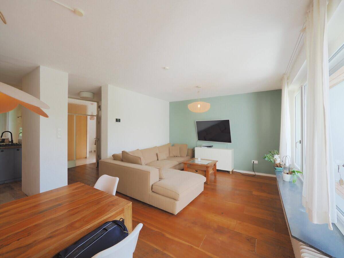 Heller Wohn- und Essbereich mit beigem Ecksofa, Holzboden und großem Fenster.