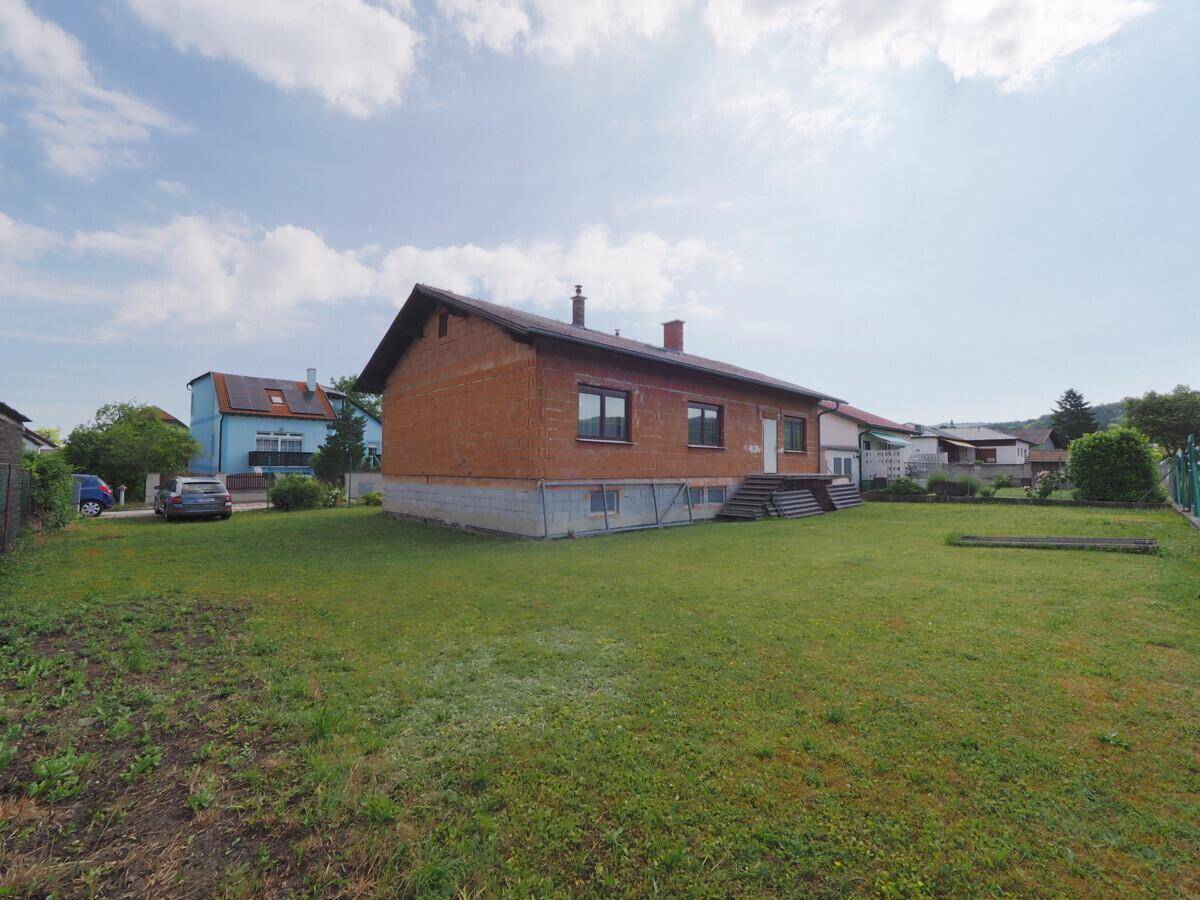 Seitenansicht des Hauses im Rohbau mit weitläufiger Rasenfläche und ländlicher Umgebung.