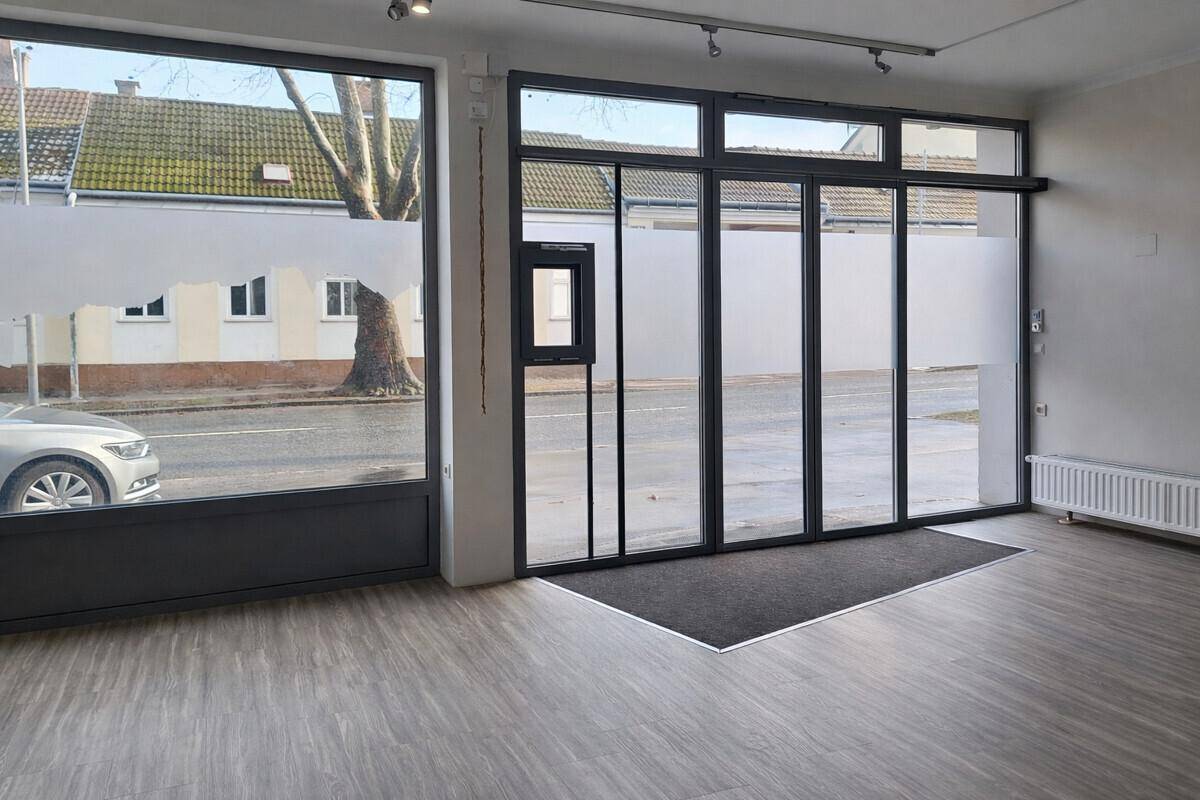 Eingangsbereich eines Geschäfts mit großen Schaufenstern und Blick auf die Straße und parkende Autos.