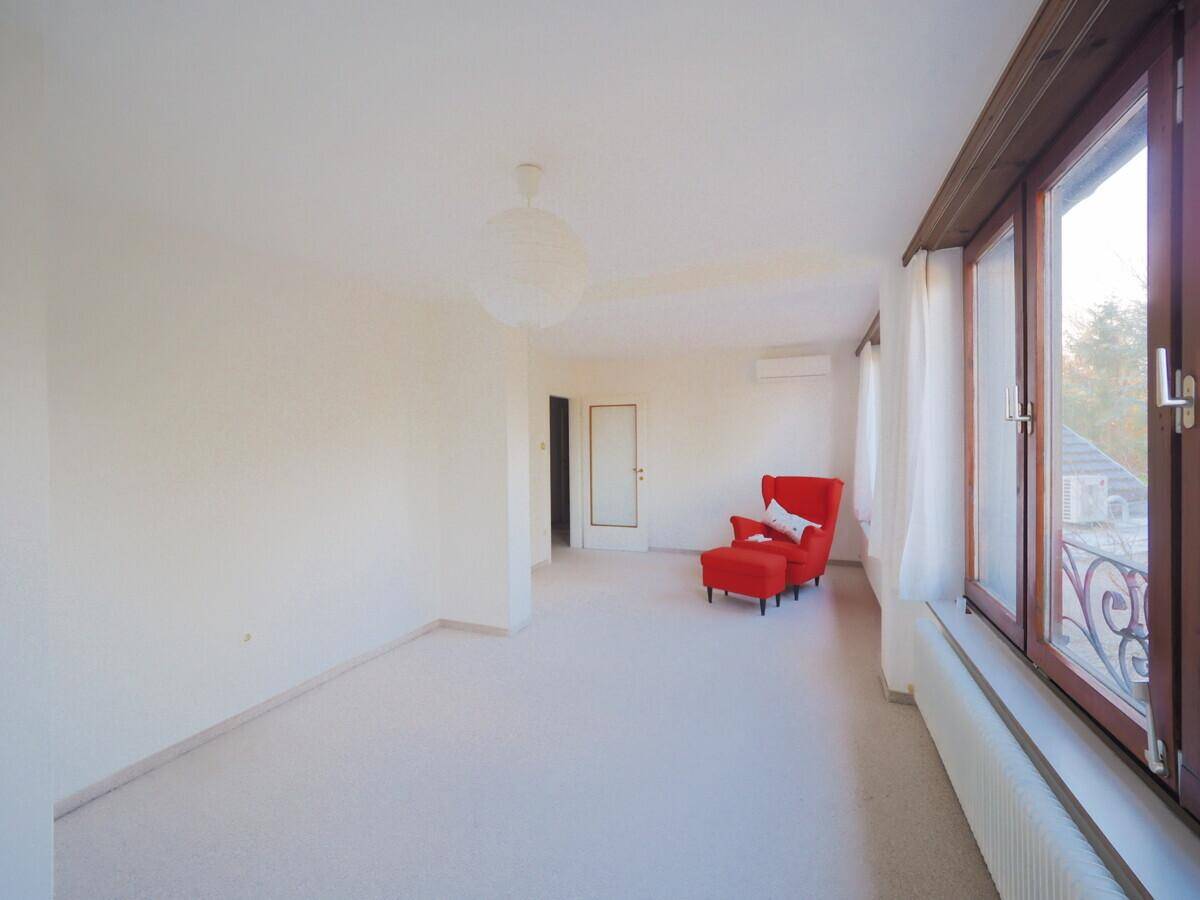 Großes, helles Zimmer mit Teppichboden, breiten Fenstern und einem roten Sessel, ideal als Wohnbereich.