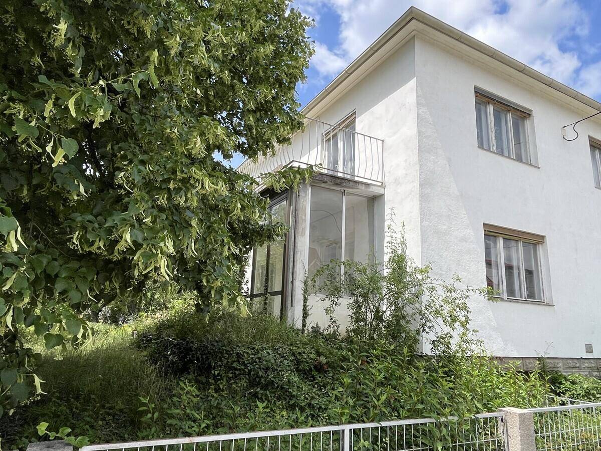 Weißes Haus mit kleinem Balkon, Wintergarten und üppiger Vegetation im Vordergrund unter blauem Himmel.