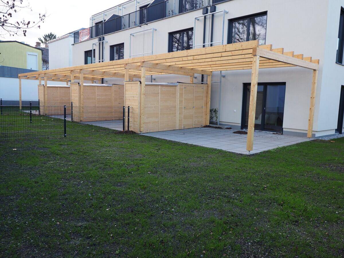 Großzügige Terrasse mit Holzpergola und angrenzendem Rasenbereich hinter dem modernen Gebäude.