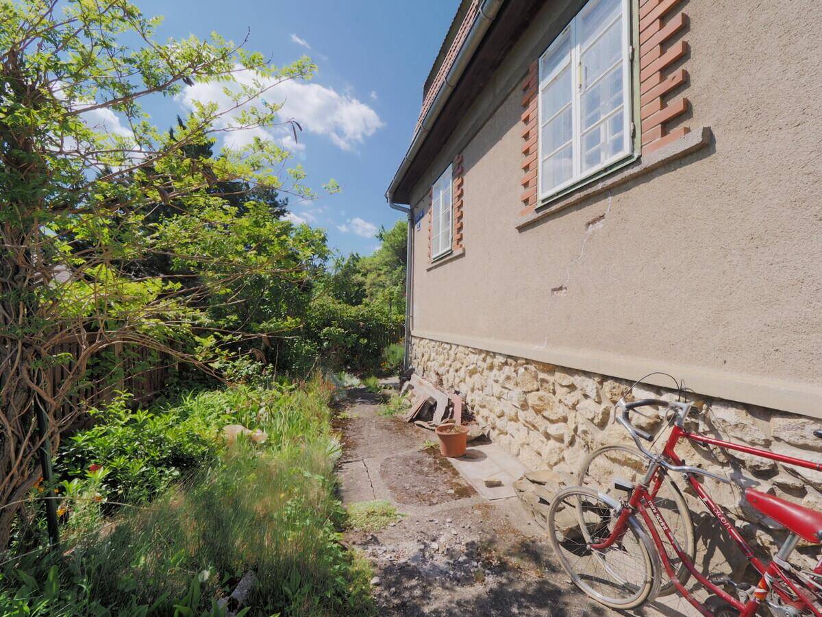 Seitenweg am Haus mit einer Steinmauer, umgeben von Vegetation und abgestellten Fahrrädern.