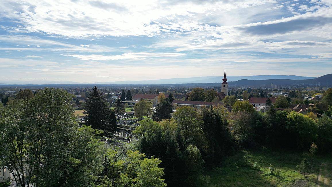 Weitläufiger Blick über eine Stadt mit Kirchturm, umgeben von Wäldern und Bergen am Horizont.