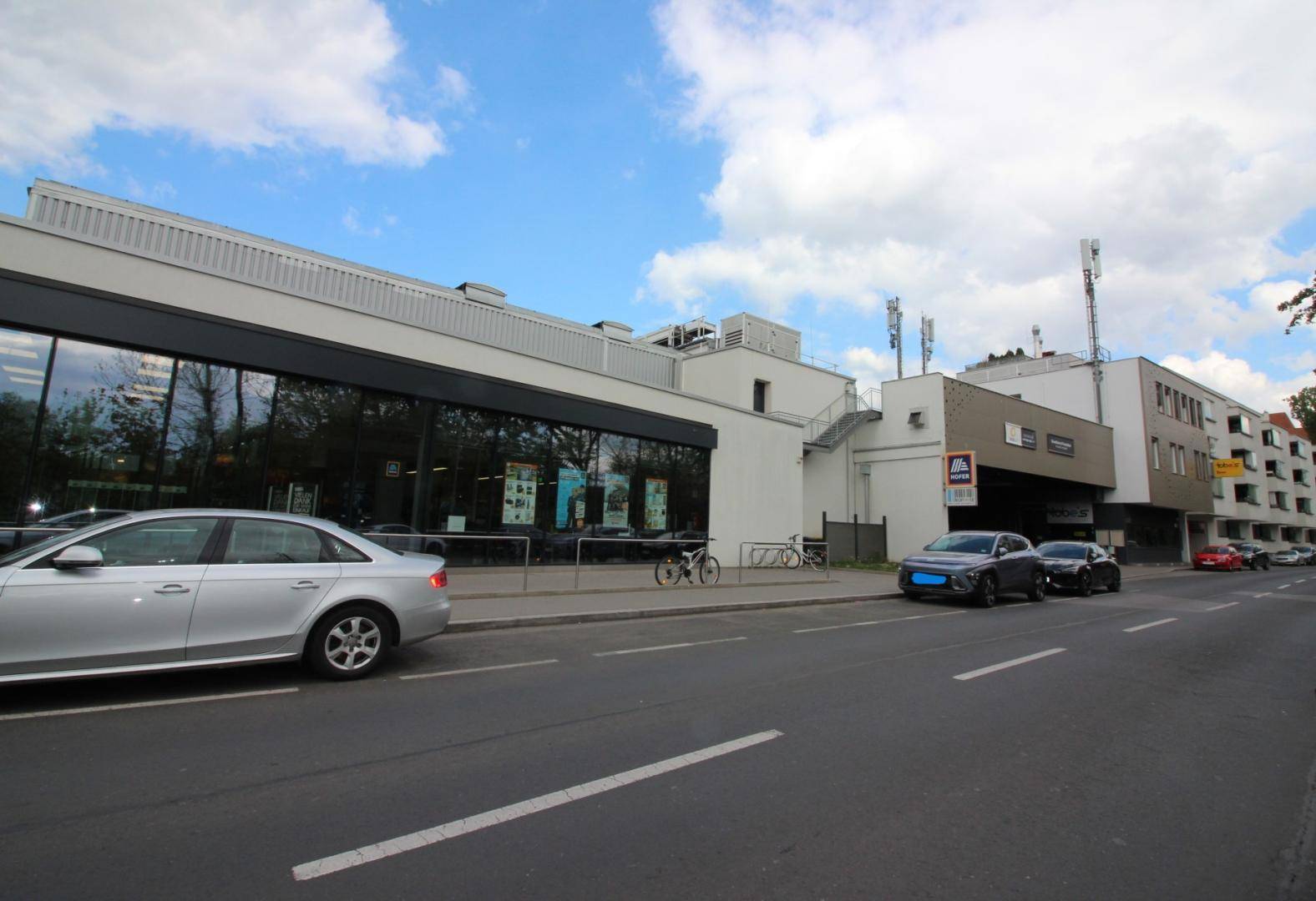 Straßenansicht eines modernen Geschäftsgebäudes mit großen Glasfronten und angrenzendem Parkplatz.