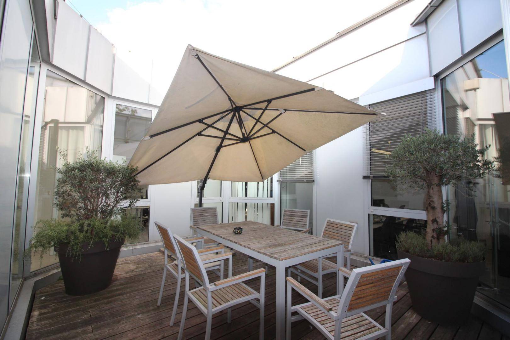 Gemütliche Dachterrasse mit Holzboden, Gartenmöbeln und Sonnenschirm, umgeben von Glasfassaden.