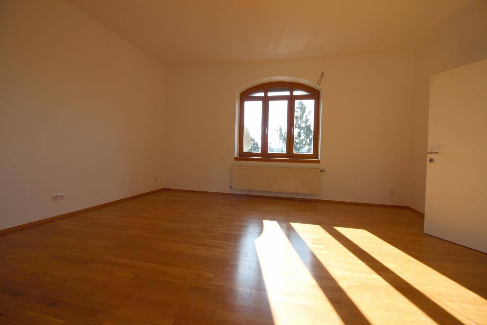 Helles, leeres Zimmer mit Holzboden, großem Fenster und viel natürlichem Lichteinfall.