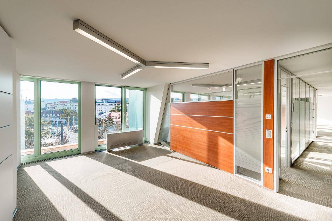 Leeres Büro mit Teppichboden, Glaswänden und großen Fenstern, die viel Tageslicht hereinlassen.