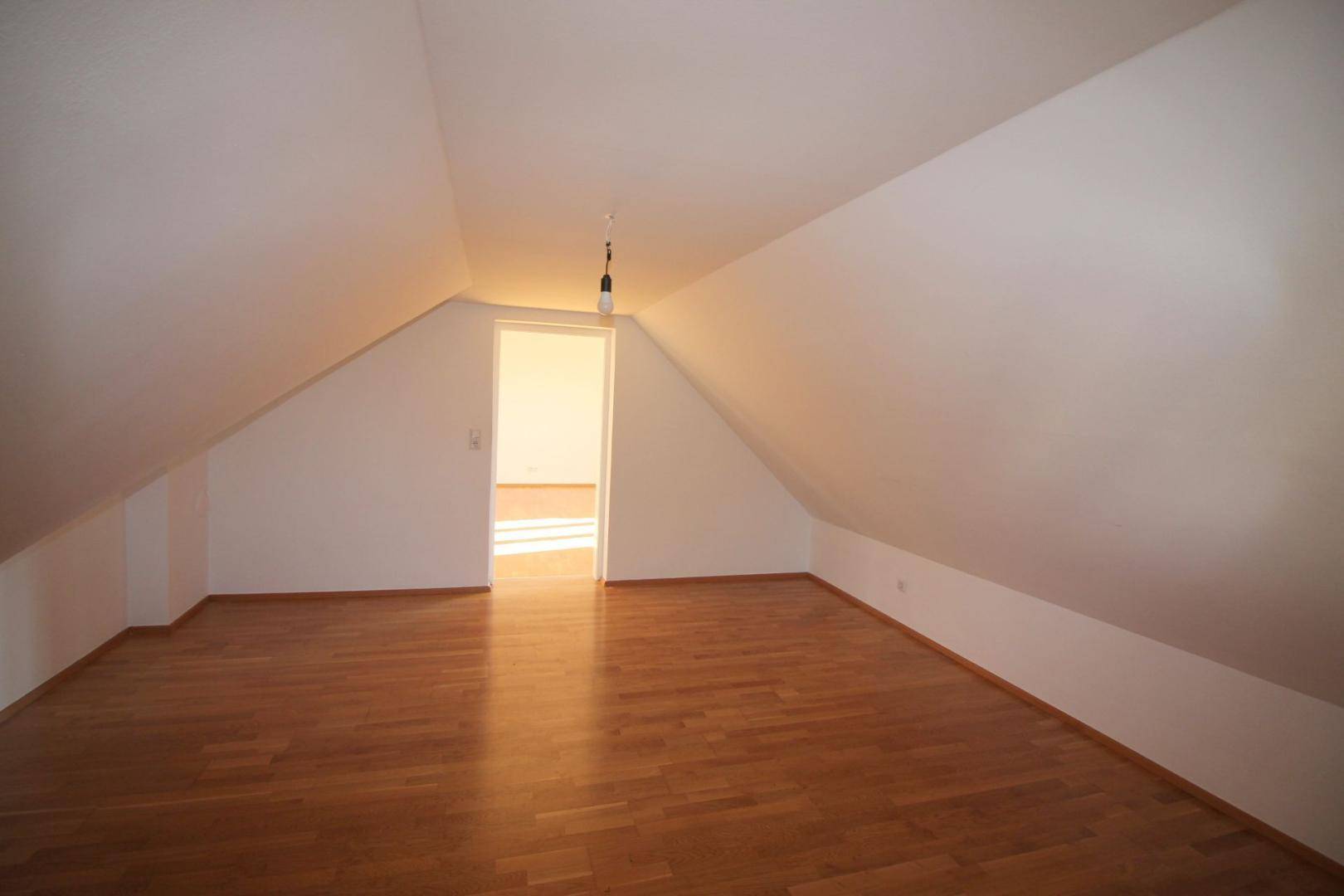 Helles Dachzimmer mit schrägen Wänden und Holzboden, das einen Zugang zu einem weiteren Raum bietet.