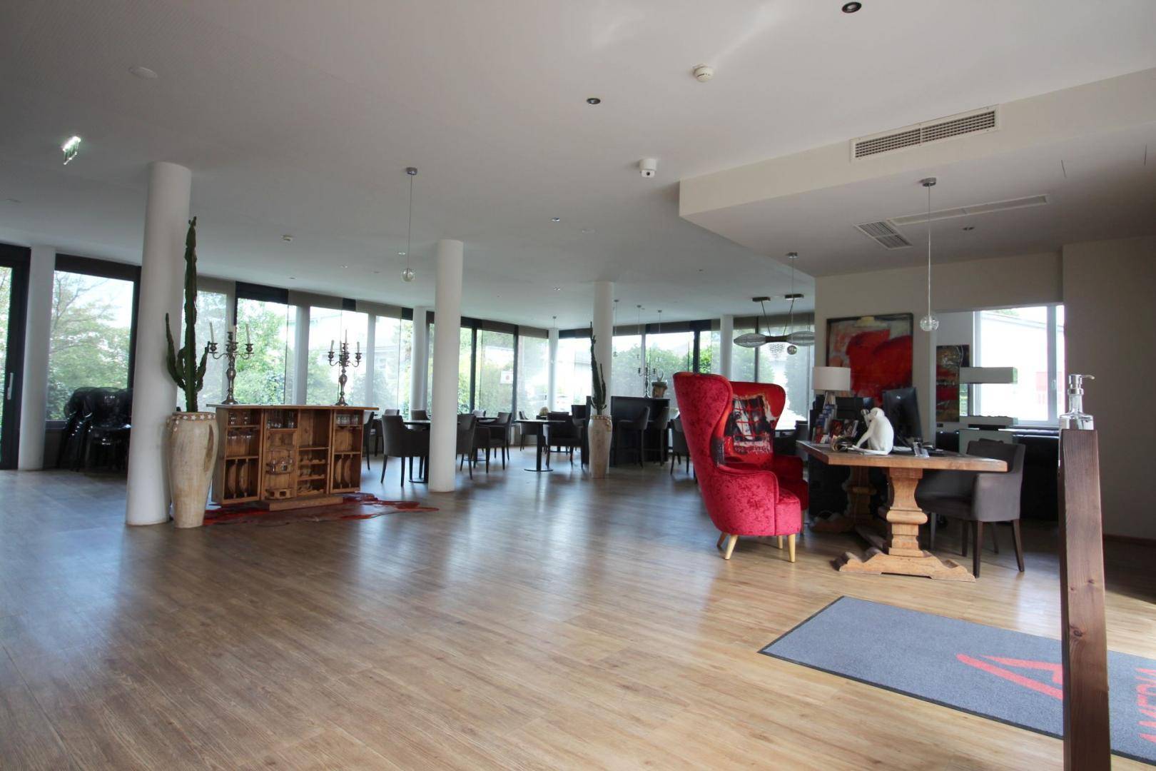 Geräumiger Loungebereich mit Holzboden, großen Fenstern und modernen Möbeln in Rot und Grau.