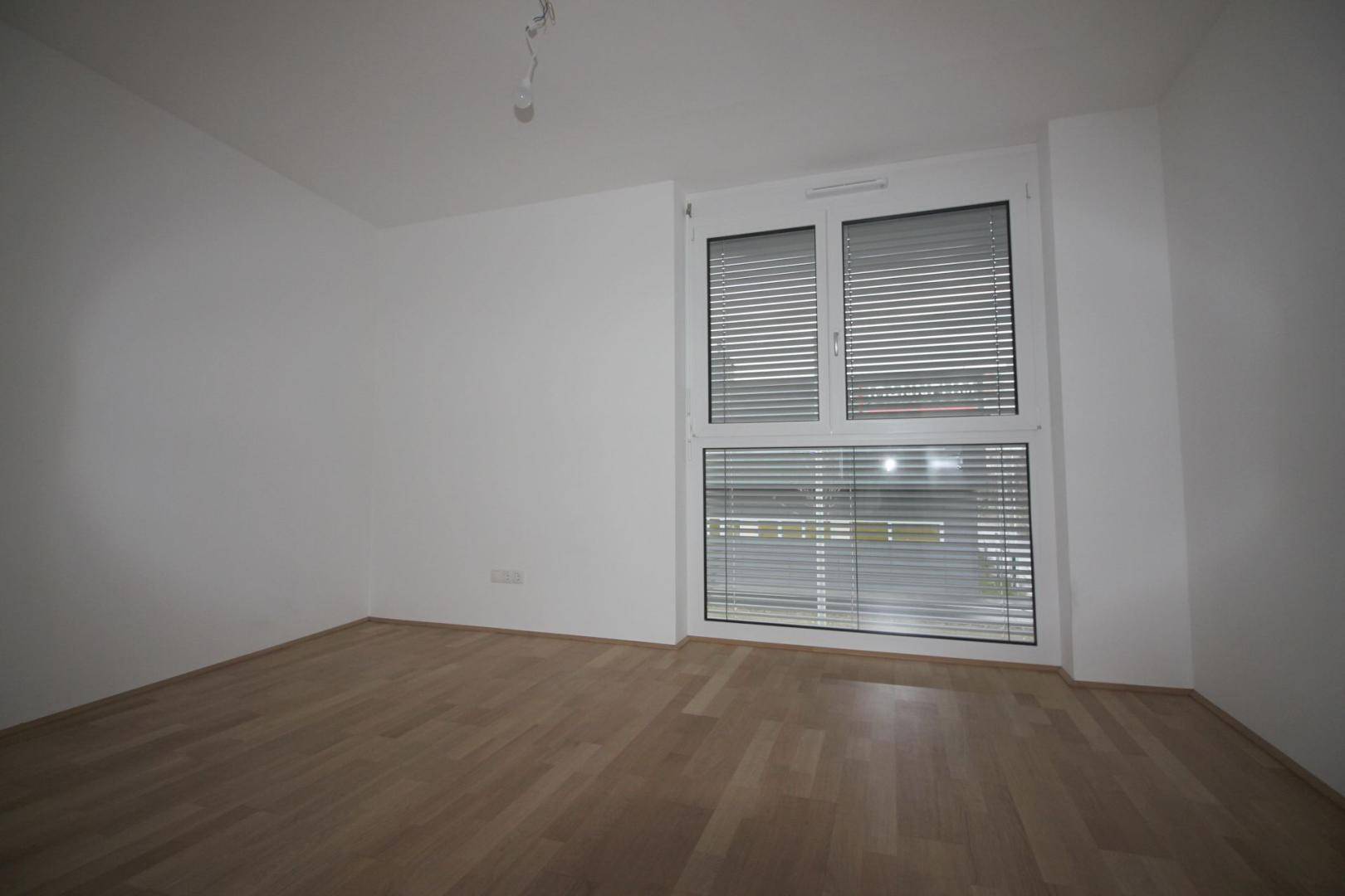 Leeres Zimmer mit Parkettboden und großen Fenstern mit Jalousien.