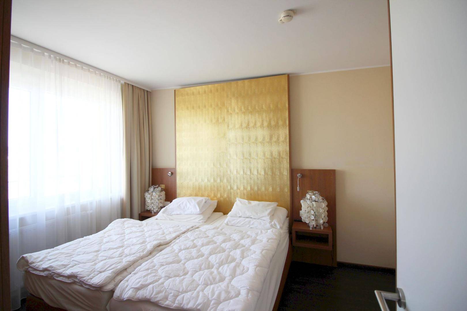 Schlafzimmer mit zwei Einzelbetten, auffälligem goldenem Kopfteil und großem Fenster für viel Licht.