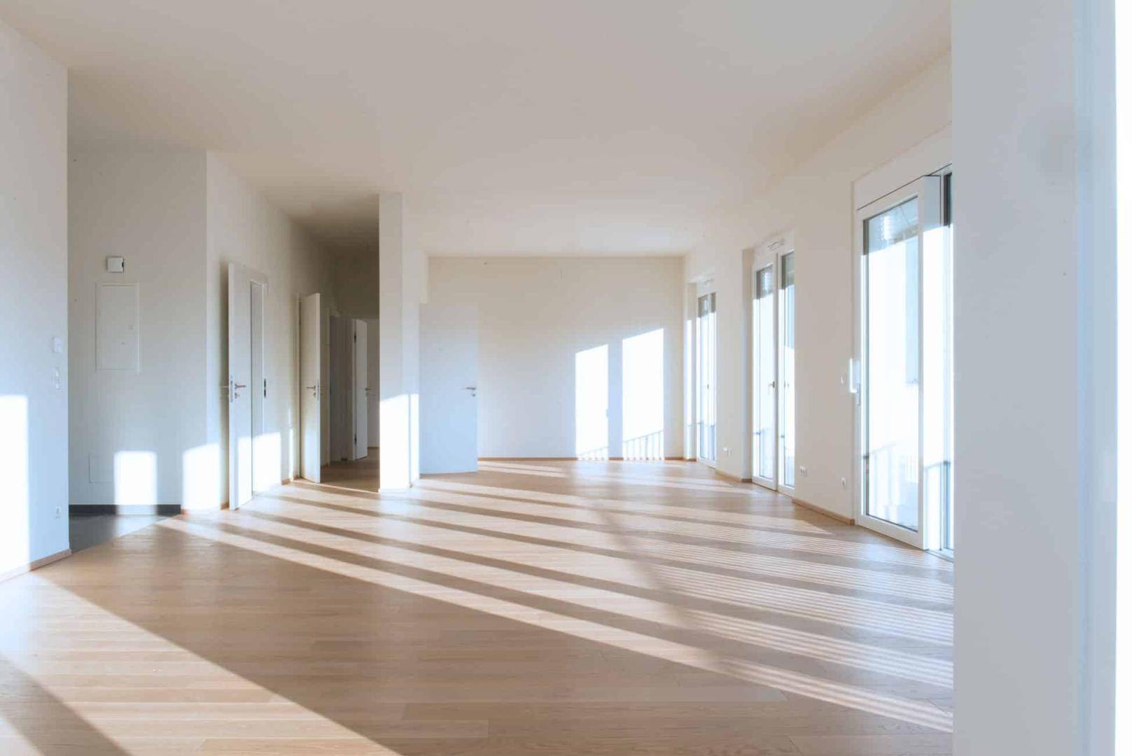 Lichtdurchfluteter Wohnbereich mit Holzboden und mehreren Fenstertüren, die viel Sonnenlicht hereinlassen und zu einem Balkon führen.