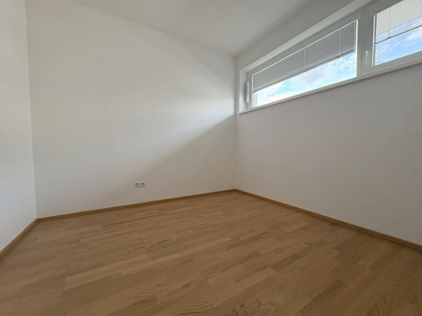 Leeres Zimmer mit hellem Holzparkettboden und einem Fenster mit Jalousien, bereit zur individuellen Gestaltung.